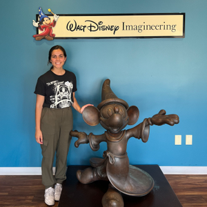 Female Disney intern with dark brown hair smiling standing next to a Sorcerer Mickey statue in front of a Walt Disney Imagineering sign